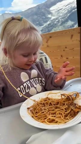 A young girl with blonde hair tied in pigtails sits in a high chair at a table and eats spaghetti. She is wearing a purple sweatshirt with the words "Cozy Vibes" printed on it. The girl is using her hands to bring the spaghetti to her mouth. In the background, there are mountains and a wooden fence.