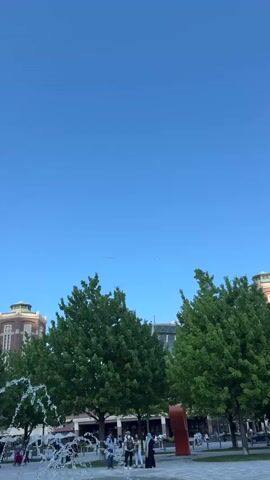 The video shows an outdoor scene with a clear blue sky, green trees, and a fountain in the foreground. Several people are walking around in the background. Above the trees, there is a small plane pulling a banner that reads "ACCIDENTS? COSTAJLANELAW.COM...1-800-800-0000!"