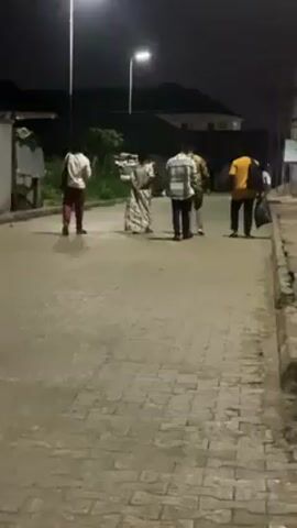 At night, five individuals are walking along a paved road illuminated by streetlights. The road appears to be lined with patterned paving stones. One person is on the left wearing a white top and maroon pants, while a woman in a light-colored dress walks alongside three other people in the center and right of the frame. These three wear different shirts, including a checkered pattern and a yellow shirt, and one carries a bag. A white car is parked on the left side of the road, and a building with multiple stories is visible in the background.