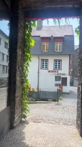 The video shows a view of a cobblestone street and buildings. A narrow stone walkway with a vine growing over it leads to an open area. In the center of the view is a two-story white building with a grey roof and dormer windows. The building has several windows with red trim. A sign on the building reads "Rose Inn." Planters with red and yellow flowers are in front of the building. To the left of the white building, another white building is visible. To the right of the white building, a narrow street curves away, with more buildings and a grey stone structure. In the distance, a blue car is parked on a cobblestone surface, and a red car is parked behind a fence. The sky is clear and blue.