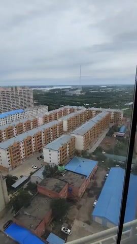 The video shows an aerial view of a residential area. In the foreground, there are several apartment buildings of varying heights and architectural styles. Some buildings have orange facades and white trim, while others are taller and feature blue and white exteriors. The streets between the buildings are visible, with cars parked along the sides. Further in the distance, more apartment complexes and some taller buildings can be seen, along with a body of water and a Ferris wheel. The sky is overcast with clouds. The camera is panning slowly to the right, offering a panoramic view of the urban landscape.