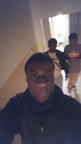 A selfie video begins with a close-up of a Black person's face, smiling. They are wearing a black jacket over a dark scarf. In the background, two other individuals are walking away down a hallway. The person on the left is wearing a yellow t-shirt and khaki shorts. The person on the right is wearing a gray t-shirt and khaki pants. The hallway appears to be indoors, with light-colored walls. The camera then moves slightly, revealing more of the hallway and the two individuals walking away. The person filming smiles again. The video continues as the person filming walks, keeping the other two individuals in view as they move down the hallway. The video ends with the person filming still smiling.