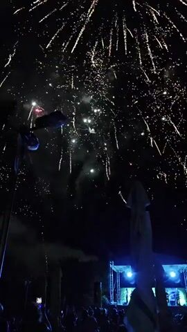 The video displays a nighttime outdoor event with fireworks illuminating the sky. A crowd of people is visible at the bottom of the frame, gathered in front of a stage where bright lights are set up. The fireworks are colorful, with bursts of pink, white, and green against the dark sky. The atmosphere appears festive, with a sense of celebration. The stage has what looks like musical equipment, suggesting a performance is or was taking place. Umbrellas are also visible in the background, possibly indicating outdoor seating or vendor stalls. The overall scene captures a dynamic event marked by light, sound, and people enjoying themselves.