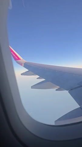 The video shows a view from an airplane window. The airplane wing is visible in the foreground, with the Wizz Air logo on the wingtip. The sky is clear and blue, with a thin layer of clouds visible on the horizon. The sun appears to be low in the sky, casting a warm glow on the wing. The video pans slightly to the right, showing more of the wing and the sky.