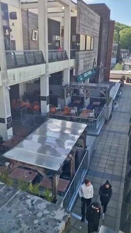 The video showcases a multi-level outdoor commercial area on a sunny day. Several people are seen walking up and down staircases, some walking as individuals, others in small groups. There are several outdoor dining areas with tables and chairs, some covered by canopies or enclosed structures. One establishment has "COFFEE - BRUNCH" and "LUNCH - DRINK" displayed. In the background, there are buildings, some under construction with scaffolding. Trees and a blue sky with scattered clouds are visible in the distance. The area is paved with a combination of square and rectangular pavers. The camera view changes frequently to capture different angles of the scene.