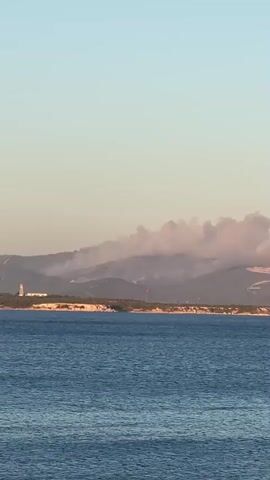 A distant view of a coastal landscape with a body of water in the foreground. On the horizon, a hill is covered in smoke, indicating a fire. A red object, possibly a flag or a marker, is visible on the hill. A white lighthouse stands on a peninsula to the left of the smoke plume. The sky is clear with a gradient of light blue to pale orange near the horizon, suggesting either sunrise or sunset. The water is a deep blue, with gentle waves. The overall scene conveys a sense of distance and potential environmental concern due to the smoke.