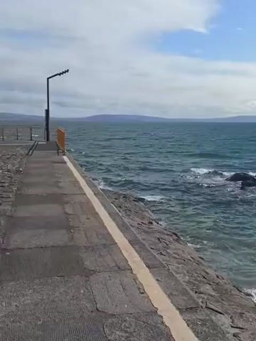 The video shows a pier extending out into a body of water. The pier is made of concrete and has a yellow line marking the edge. To the right of the pier, there is a rocky shoreline. The water is choppy and there are waves breaking against the rocks. In the distance, there is a landmass, likely an island or coastline, under a partly cloudy sky. A single lamppost is visible on the left side of the pier. There are no people or animals visible in the video. The video appears to be shot from a stationary position, looking out towards the water.