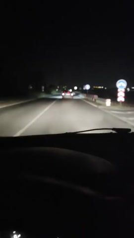 A dark night scene from inside a vehicle looking forward. The headlights of the vehicle illuminate the road ahead, showing a white line on the left and a guardrail on the right. Another car is visible in front, its taillights glowing red. The car's license plate reads "B 6711TT". The scene suggests driving on a road at night, possibly a highway, with limited visibility beyond the illuminated area.