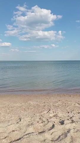 The video shows a view of a sandy beach meeting the ocean under a bright blue sky with scattered white clouds. The camera is positioned low, looking out towards the water. The sand in the foreground is light brown and appears dry with some texture from footprints. A thin line of wet sand is visible where the water meets the shore. The ocean water is a calm, clear blue-green. In the distance, the horizon separates the water from the sky. White, puffy clouds drift across the sky.