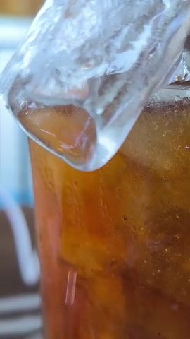 The video shows a close-up shot of a glass filled with iced tea. A large ice cube sits at the top of the glass. Condensation is visible on the exterior of the glass. The tea is a light brown color.  In the background, a blurred white object and brown surface can be seen.