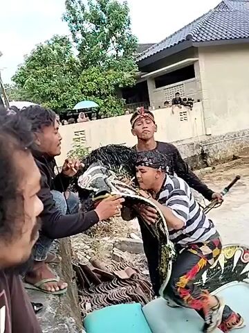 The video shows a group of people outdoors. In the foreground, the back of a person with long, curly dark hair is visible. To their right and slightly behind, two young men are interacting. One, wearing a black long-sleeved shirt and jeans, is crouching. The other, in a black long-sleeved shirt and headscarf, is standing and appears to be facing the crouching person. Another person in a black jacket with white detailing is standing to the left, partially obscuring the view of the two young men. A person holding a green and yellow umbrella stands behind them. In the background, there are more people, trees, and structures that suggest an outdoor gathering or event. The ground appears to be uneven and somewhat muddy.