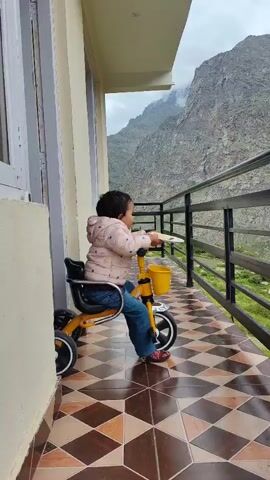 A toddler is sitting on a yellow tricycle on a balcony. The toddler is wearing a pink jacket and blue jeans. The tricycle has a yellow basket on the front. The balcony has a tiled floor and a railing. In the background, there is a mountain and a cloudy sky. The video is from a side view, showing the toddler from the waist down.