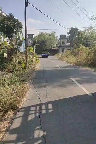 The video shows a bright, sunny day with a clear blue sky. A paved road stretches into the distance, flanked by dry, yellowish vegetation and some trees. On either side of the road, near the entrance of what appears to be an estate or property, are two weathered, square stone structures with arched openings and small windows at the top. A small blue truck, laden with a stack of round, light-colored objects, possibly coconuts or melons, is driving away from the camera towards these structures. The truck is positioned centrally on the road, heading towards the gate-like entrance. The overall scene suggests a rural or semi-rural setting.
