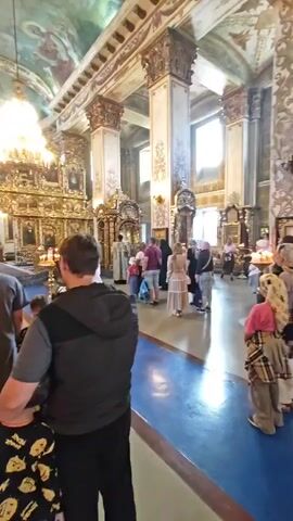 The video opens with a view of the interior of a church. A man in a grey t-shirt and dark vest is in the foreground, facing away from the camera. He has short brown hair. To his left, a woman with dark hair tied back is also facing away from the camera. Behind them, several people are gathered, some wearing headscarves. The church interior is ornately decorated with gold accents and religious imagery on the walls and ceiling. A large chandelier hangs from the ceiling. In the background, a priest in ceremonial robes stands near an altar. Other individuals, including children, are visible walking and standing within the church. The floor is a polished blue. The camera pans slightly to the left, showing more of the church's interior, including tall, decorative columns and more religious artwork. The video continues with a similar view, focusing on the altar area and the people present. The overall impression is of a religious service or a visit to a historical church.