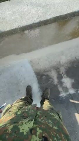 The video shows a person wearing military fatigues standing on a concrete ledge, possibly the roof of a building. The camera pans up and outward, revealing a view of a building across a courtyard with trees and a parked car.  Two people are visible walking toward the building. The sky is clear and blue.  The ledge the person stands on is partially covered in a white, powdery substance, possibly snow or residue. The overall setting appears to be an outdoor urban environment.