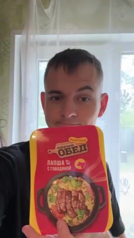 A man stands in front of a window, holding up a package of “Rollton” instant beef noodles. The package is red and yellow, with a picture of noodles and beef on the front. The man is wearing a black t-shirt with a white lightning bolt logo. He looks at the camera and smiles. The background includes a window, a white curtain, and green patterned tile on the wall.