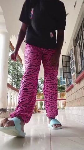 A young woman in a black t-shirt, pink patterned pants, and blue sandals is doing push-ups on a tiled porch. She also wears a red satin bonnet with a gold pattern. The porch has white columns and a white railing. There is a window behind her and a small dog lying down in the background. The woman smiles at the camera as she starts the push-up.