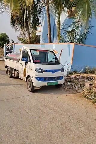 The video shows a parked, white, electric pickup truck with a tank mounted on its bed. The tank is labeled “Shirdi Sai Drinking Water” and has a phone number printed on it. The truck is parked on a paved road next to a blue building. There are trees in the background. The truck appears slightly dirty. The video seems to be taken on a sunny day.