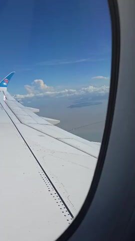 The video shows a view from an airplane window. The left side of the frame is dominated by the white wing of the airplane, which extends towards the right edge. The wing has a series of small dots or rivets running along its edge. The tip of the wing is blue and features a logo, possibly an airline logo. Beyond the wing, the sky is a clear blue, with a few white clouds scattered across it. Below the clouds, a hazy landscape or body of water is visible, with a long, thin, dark line extending across it, potentially a bridge or a ship. The right side of the frame is occupied by the dark circular frame of the airplane window.