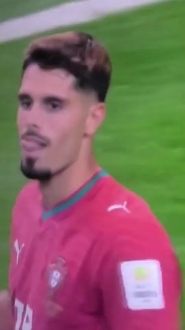 The video shows a male soccer player in a pink jersey with teal trim and white accents. He has short, light brown hair styled with darker roots. The player is looking off to the side with his mouth slightly open. In the background, other players in red and white jerseys are on a green soccer field with white markings. There are spectators in the stands and various advertisements around the field. A logo with "FIFA" in large yellow letters briefly appears over the player's arm.