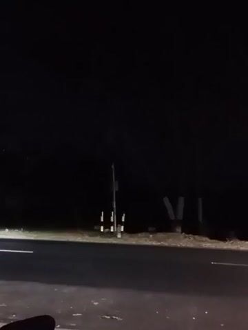 The video begins by showing a nighttime scene of a street with a single streetlamp in the center of the frame. The streetlamp is illuminated and casts a warm glow. To the left of the streetlamp, there is a white line painted on the road, indicating a lane marker. The road itself appears to be asphalt. Behind the streetlamp, there are trees and vegetation, which are mostly obscured by darkness. There are also some utility poles visible in the background. The camera pans slightly to the right, revealing more of the dark surroundings and the trees. The video continues to show the streetlamp and its surroundings as the camera moves. In the latter half of the video, the streetlamp appears to flicker and dim, and the overall scene becomes darker as if the light source is weakening or being obstructed. The video ends with the streetlamp barely visible and the surroundings completely dark.