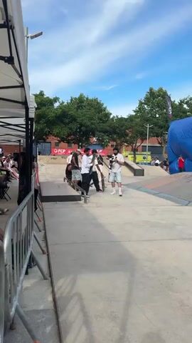 A video shows a group of men in a skatepark on a sunny day. A man in a white long-sleeved shirt and black pants approaches the camera, holding a skateboard. Another man in a white short-sleeved shirt and black pants walks past him, also holding a skateboard. A man in a white t-shirt and light blue shorts films them with a professional camera. Other men are visible in the background, some on skateboards. The skatepark features ramps and rails, and a large blue tarp structure stands on the right. Green trees and a blue sky with clouds are in the background.
