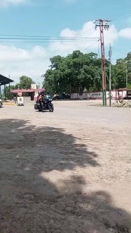 The video shows three motorcycles driving across a dirt area. The sky is blue with some clouds. There are buildings, trees, and utility poles in the background. The first motorcycle is a black touring bike with two people on it. The second is a blue touring bike with two people on it. The third is a red motorcycle with one person on it.
