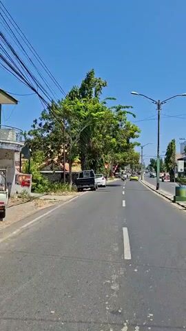 The video shows a road with vehicles and trees. The road is paved with asphalt and has white lane markings. There are several vehicles on the road, including a black pickup truck, a white car, and other vehicles in the distance. Trees are present on both sides of the road, with lush green foliage. The sky is clear and blue. The scene appears to be a daytime shot in a populated area, possibly a town or city. There are also power lines visible above the trees.