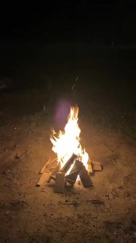 The video shows a campfire burning in the dark. The fire is in the center of the frame, with flames reaching upwards. The ground around the fire is dirt and grass. There are pieces of wood arranged in a pyramid shape to form the base of the fire. The background is dark and out of focus, suggesting it is nighttime. There are no people or animals visible in the video. The fire appears to be safely contained within a designated area.