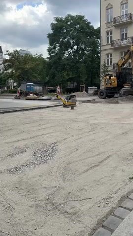 A construction site on a cloudy day. The foreground is covered in light-colored gravel and dirt, with tire tracks visible. In the middle ground, a small yellow excavator is parked on the gravel. Further back, a larger yellow excavator is operating on the right side of the frame, its arm extended. To the left, a blue truck with a white tank sits behind a barrier. A white plate-like object is partially visible on the ground near the truck. A tall, multi-story building with tan walls and white trim stands in the background on the right. Trees are visible behind the building and on the left side of the frame. The ground is being compacted with a piece of construction equipment that appears to be a plate compactor. The overall scene suggests road or pavement construction is underway.