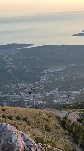 The video shows a scenic view from a mountaintop overlooking a bay and a town. A cable car system is visible, with several red gondolas suspended in the air. The gondolas are moving along a cable stretched between towers. The town below appears to be nestled in a valley, with buildings and roads spread out. The bay is calm and reflects the soft light of the sky, which suggests it is either sunrise or sunset. The surrounding hills are covered with green vegetation, and there are some rocky outcrops in the foreground. The overall atmosphere is peaceful and serene.