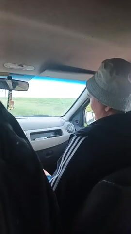 A person wearing a hat and a black jacket with white stripes on the sleeves is seated in the driver's seat of a car. They are adjusting their hat. The interior of the car is visible, including the dashboard, rearview mirror, and ceiling. Outside the car, a green field is visible through the windshield.
