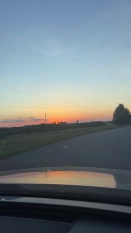 This video depicts a scenic drive through a rural landscape in Dovhe, Ukraine, during sunset. The camera is positioned inside a vehicle, looking out through the windshield. The foreground shows the car's dashboard and the reflection of the sky and landscape on the windshield. The main subjects are the natural scenery and the road.

The setting is a wide-open, grassy area with scattered trees and bushes. A paved road curves to the right, leading into the distance. The sky is a beautiful gradient of soft blues, oranges, and yellows, indicating the sun is setting. The sun itself is partially visible on the horizon, casting a warm glow. The overall mood is peaceful and serene, evoking a sense of tranquility associated with a sunset drive. There is no visible text in the video. The time of day is clearly sunset. The weather appears to be clear and pleasant, with a gentle breeze suggested by the way the trees are standing. The location cues, such as the road and the type of vegetation, are consistent with a rural setting in Ukraine.