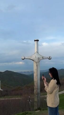 The video presents a scenic view featuring three large, silver sword monuments, set against a backdrop of rolling green hills and a partly cloudy sky. A woman with dark hair, wearing a tan jacket and jeans, is seen standing to the right of the frame, holding a cell phone and appearing to take a photo of the sword monument. The monuments themselves are positioned on a grassy hilltop, overlooking a landscape with winding roads visible through the trees. The camera pans slightly, offering a broader view of the environment, before returning to the central image of the swords. The overall setting suggests a rural, possibly mountainous region, with a touch of historical or memorial significance implied by the presence of the sword monuments.