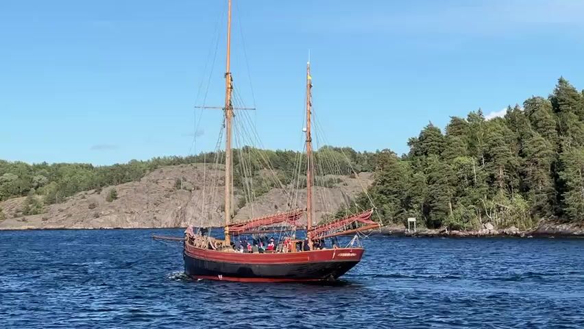 A wooden schooner with two masts and red sails is sailing on dark blue water under a clear blue sky. The schooner is dark red and black with a wooden deck. There are several people on the deck, some standing and some seated. The boat is moving towards the right side of the frame. In the background, there is a rocky, tree-covered island on the left and a forested island on the right. The water is slightly choppy. A white marker buoy is visible in the water near the right island.