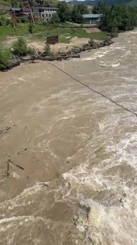 The video shows a murky, fast-flowing river with debris such as logs and trash floating on the surface. The water is turbulent, with white rapids and churning eddies. On the left bank of the river, there is a concrete structure and trees. In the background, there are buildings and a road. The perspective is from above, looking down at the river. There are no visible people in the frame. A black cable stretches across the river diagonally.