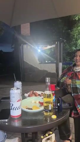 The video begins with a close-up of a round glass-top table at night. On the table are two cans of Budweiser beer, one open with a straw, a bottle of Corona beer, a plate of food that appears to be sausages and other appetizers, a phone, and a red spray can. The camera then pans to show a woman sitting in a chair to the right, wearing a plaid shirt and jeans, and a blanket over her shoulders. She is smiling and looking away from the camera. The background shows an outdoor patio area with a brick structure and some trees. The lighting suggests it is evening or nighttime. The camera then shifts focus to another person, partially visible, also seated at the table. The scene appears to be a casual gathering.