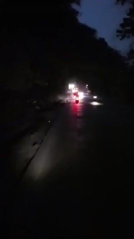 The video is shot from the perspective of someone on a motorcycle at night, looking forward. The road ahead is wet and illuminated by headlights from oncoming vehicles and the motorcycle's own light. Several cars and at least one motorcycle are visible, their lights creating streaks and reflections on the wet surface. Trees and vegetation line the sides of the road, obscured by darkness. The overall atmosphere is dark and somewhat hazy, likely due to moisture in the air or on the road. The footage is shaky, indicating movement.
