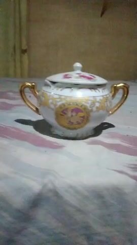 A white porcelain sugar bowl with gold handles and a floral lid sits on a patterned surface. The bowl has ornate gold and pink detailing. The background is out of focus and appears to be a wall.