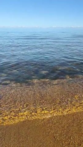 This is a vertical, eye-level, static shot of a calm body of water with a sandy shore in the foreground. The water is clear and shallow near the shore, revealing a golden-brown sandy bottom with small pebbles. Gentle waves lap at the shore. In the distance, the water transitions to a deeper blue, and a hazy, light blue sky is visible above. A faint line of white, possibly snow-capped mountains, is discernible on the horizon where the water meets the sky. There are no people or man-made objects in the frame. The overall impression is one of natural tranquility.