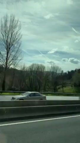 The video shows a view from a moving vehicle on a highway. Other vehicles, including cars and a tour bus, can be seen driving in the opposite direction. The sky is partly cloudy with contrails visible. The landscape beside the highway features trees and green hills. There's a concrete barrier between the lanes.