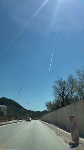 The video depicts a car driving on a road under a bright blue sky with a contrail visible above. The road has yellow lane markings, and there are street lights along the side. A gray concrete wall runs along the right side of the road, with trees visible above it. In the distance, there are green mountains. Several cars are visible on the road ahead. The video appears to be shot from the driver's perspective.