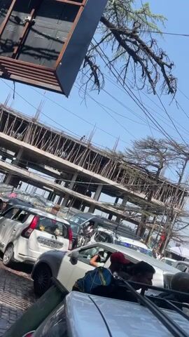 The video shows a busy street scene with heavy traffic congestion. Numerous cars are stalled, creating a long queue. In the background, a multi-story building is under construction, with exposed rebar and scaffolding visible. Another building to the right is older, with several signs for businesses like "HOTEL," "PLAZA HOTEL," and "TONI'S PIZZA WRAPS SHAKES." The street is also cluttered with a dense network of overhead electrical wires. People are visible walking and standing around the vehicles, some appearing to be drivers or pedestrians navigating the crowded area. The overall impression is one of a bustling urban environment with ongoing construction and significant traffic.