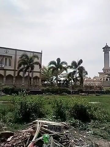 The video presents an outdoor scene under an overcast sky. It begins by showcasing a two-story building on the left, characterized by its cream-colored facade and multiple arched windows. Several palm trees stand in front of this structure. The foreground is dominated by a lush green field, appearing somewhat wild with various weeds and plants. As the camera slowly pans to the right, the field extends across the frame. In the distance, a tall, slender minaret-like tower with a domed top comes into view, flanked by other buildings that appear to be part of a larger complex, likely a mosque or religious center. The architecture suggests an Islamic influence, common in Indonesia. The scene is devoid of people or specific activities, focusing instead on the landscape and structures. The time of day is daytime, evidenced by the natural light, despite the cloudy conditions. No visible text is present. The setting is consistent with the provided location context of Kruengmate, Indonesia.