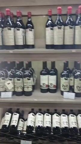 The video shows a well-lit display of wine bottles in a store. The bottles are arranged on wooden shelves and in racks. The first shelf displays several bottles of red wine with red caps. The second shelf holds a variety of wine bottles, including some with gold caps, some with purple labels, and some with orange and pink hues, possibly indicating different wine types or vintages. The third shelf contains more red wine bottles. A rack in the foreground displays more wine bottles, some with white labels. To the left, a tall bottle of liquor with a gold cap stands out. The background appears to be a store interior, with a window visible on the right side that looks out onto an outdoor area with greenery. The lighting is consistent throughout the scene. There are no people visible in the video.