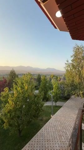 The video shows a view from a balcony or patio overlooking a park-like area with trees, grass, and distant mountains. The sun is setting, casting a warm glow on the landscape. The camera pans slightly to the left, capturing the full panorama. The scene appears peaceful and serene.