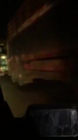 A vehicle is traveling down a dark street, passing several large trucks parked on the side of the road. The trucks are predominantly green and yellow. Streetlights and headlights are visible in the distance. The perspective is from inside the moving vehicle.