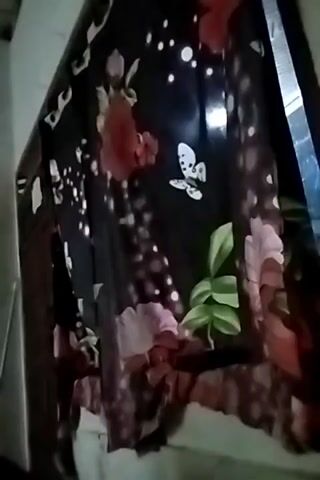 The video shows a close-up of a black fabric with a floral pattern. The fabric is draped over something, possibly a window or a piece of furniture. The pattern includes large pink and red flowers, green leaves, and small white circles that resemble bubbles or light spots. There is also a white butterfly with black spots depicted on the fabric. The lighting is dim, and the fabric appears to be slightly wrinkled. The background is blurred and out of focus, suggesting it is not the main subject of the video.