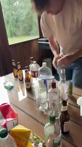 The video shows a group of people preparing a drink at a table laden with alcoholic beverages, including various liquor bottles, beer bottles, and mixers. One person is pouring a green and white container of liquid into a large, clear plastic container filled with an orange-colored liquid. There are bags of chips and boxes of juice also on the table. The event appears to be taking place outdoors on a porch or deck, with trees visible through the window. The person pouring mentions the liquid is leftover from his sister's party.