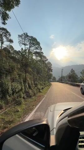 The video captures a scenic drive through a mountainous region, likely in Dharamshala, India, during sunrise. The camera is positioned inside a moving vehicle, showing a portion of the car's dashboard and side mirror. The road winds through lush green hillsides lined with pine trees. A white van is seen driving ahead on the road. As the vehicle ascends, the landscape opens up to reveal a misty valley with a town or settlement nestled on the mountainside. The sun is rising, casting a warm glow and creating sunbeams through the hazy atmosphere. The overall mood is tranquil and serene, evoking a sense of journey and natural beauty. No visible text is present in the video.